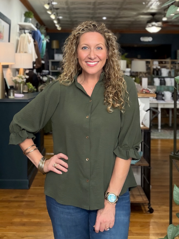 A smiling woman with curly blonde hair stands in a retail store wearing an olive green button-up blouse with ruffled three-quarter sleeves and blue jeans.