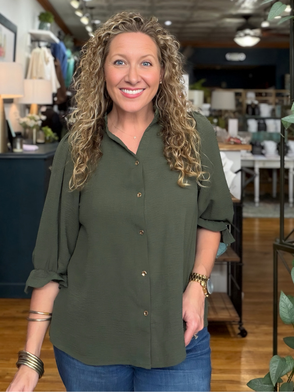 A smiling woman with long curly blonde hair wearing an olive green button-down shirt and blue jeans, standing inside a retail store.