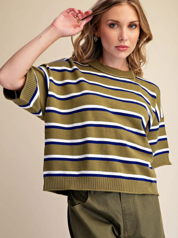 A woman wearing a cropped short-sleeve knit sweater with horizontal olive green, white, and navy blue stripes, paired with olive green pants, against a solid beige background.