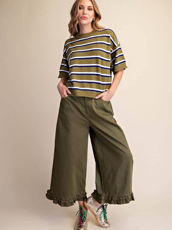 A woman modeling a short-sleeve olive green knit top with navy and white horizontal stripes, paired with wide-leg olive green pants featuring ruffled hems, against a solid beige studio background.