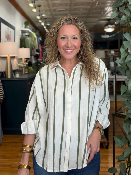 A smiling woman with curly blonde hair wearing a cream-colored linen button-up shirt with vertical olive green stripes, standing in a retail store.