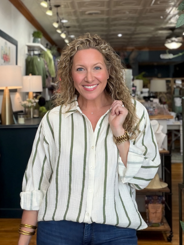 A smiling woman with curly blonde hair stands in a boutique wearing a white button-up shirt with vertical sage green stripes and dark blue jeans.