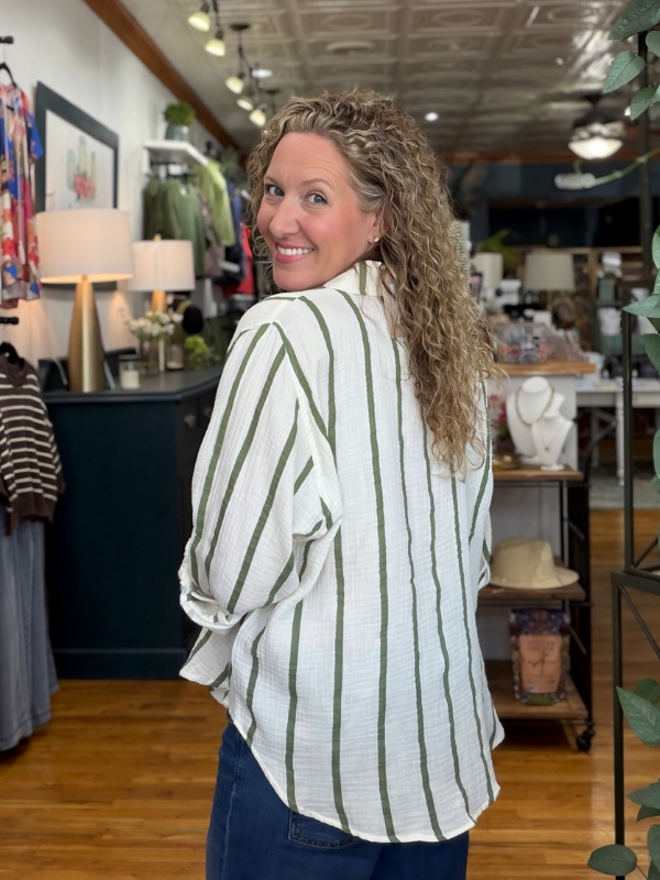 A woman with curly hair wearing a white and green vertically striped button-up shirt and dark blue jeans, standing inside a retail boutique.