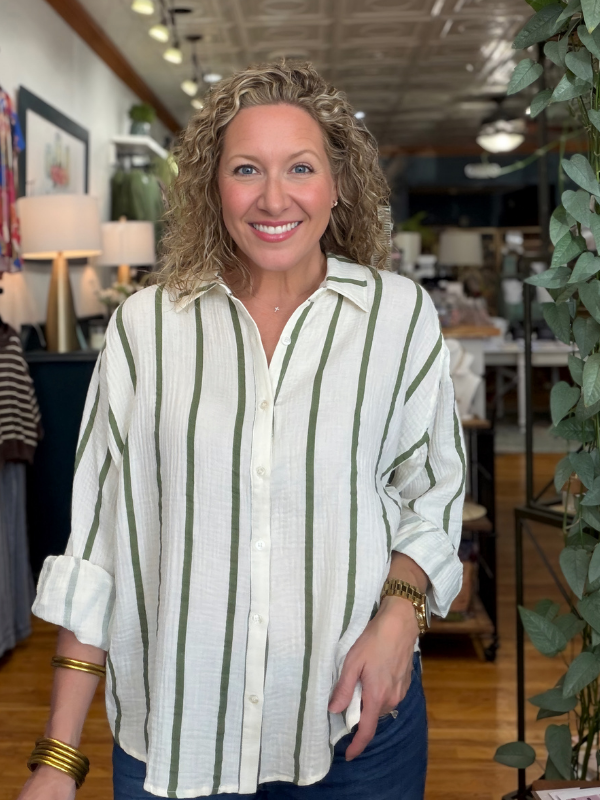 A smiling woman with curly blonde hair wearing a white linen button-up shirt with vertical sage green stripes, standing inside a boutique.