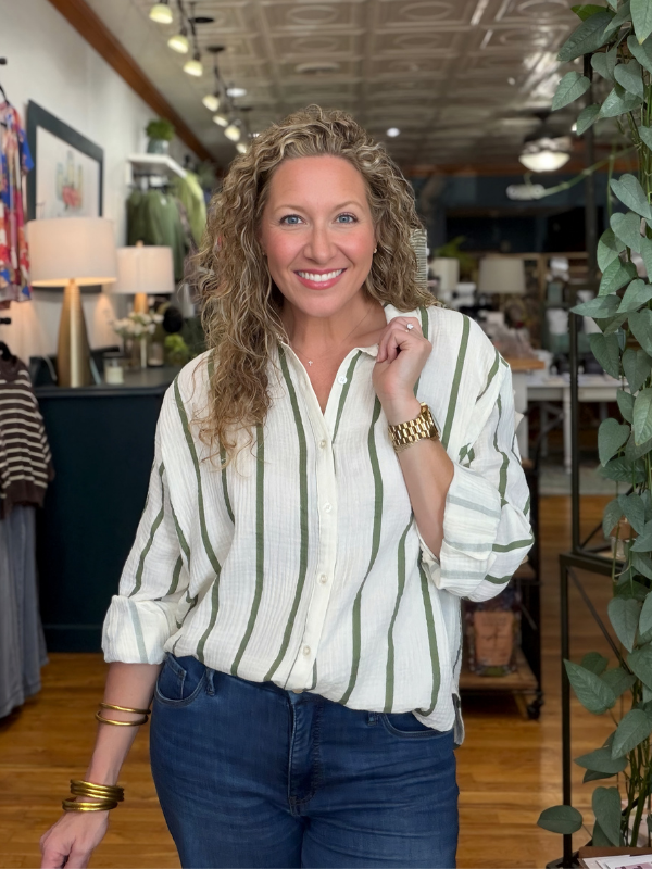 A smiling woman with curly blonde hair stands in a boutique wearing a cream-colored button-down shirt with vertical green stripes and dark blue jeans.