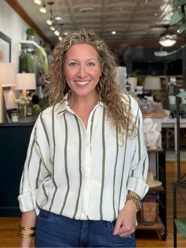 A smiling woman with curly blonde hair wearing a white button-up shirt with vertical olive green stripes and blue jeans, standing in a well-lit home decor or furniture store.