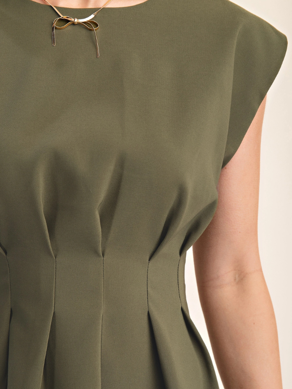 Close-up of a person wearing an olive green sleeveless dress with a delicate gold bow necklace at the neckline, set against a plain light background.