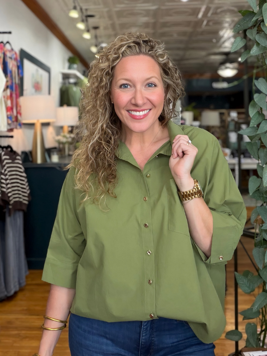 A smiling woman with curly blonde hair wearing an olive green button-down shirt and blue jeans, standing in a retail store interior.