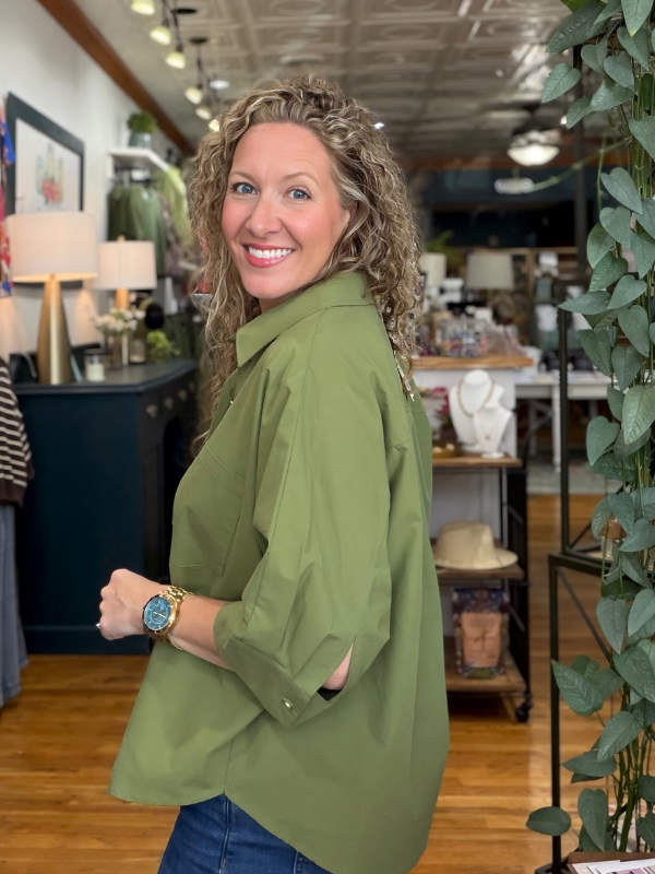 A smiling woman with curly blonde hair wearing an oversized olive green button-down shirt and blue jeans, standing inside a retail store with wooden floors and shelves in the background.