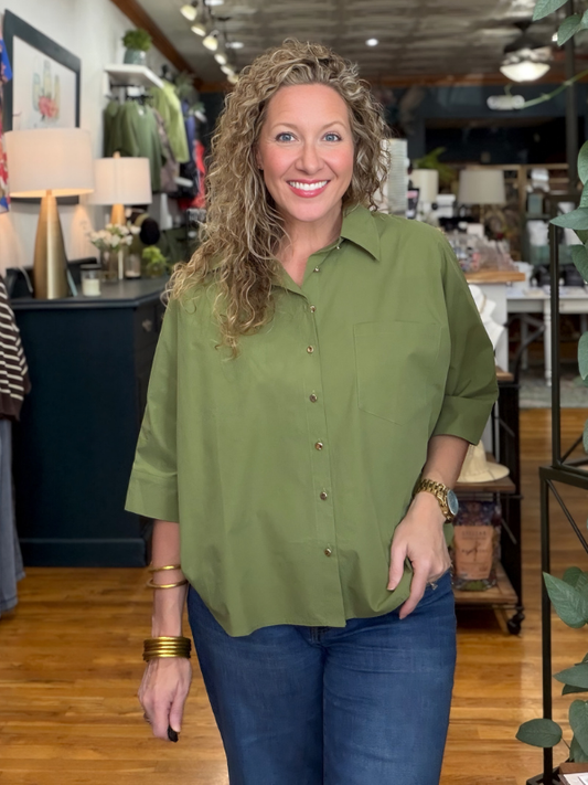 A smiling woman with curly blonde hair wearing an olive green button-down shirt and blue jeans, standing inside a retail store.