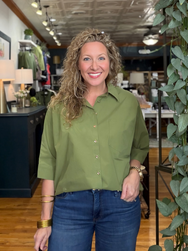 A smiling woman with curly blonde hair stands in a retail store wearing an olive green button-down shirt and dark blue jeans.