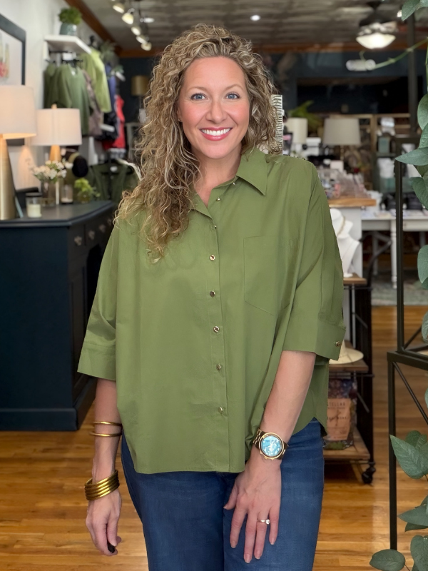 A smiling woman with curly blonde hair wearing an olive green short-sleeve button-up shirt and blue jeans, standing inside a retail store.