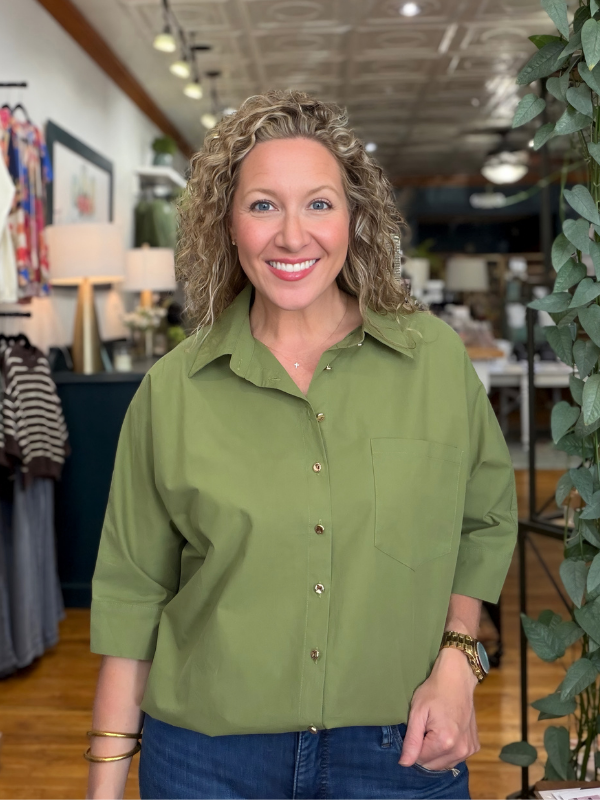 A smiling woman with curly blonde hair wearing an olive green button-down shirt and blue jeans, standing in a retail store interior with plants and clothing racks visible in the background.