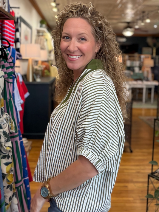 Woman in a striped shirt standing in a store with clothing racks and products in the background.