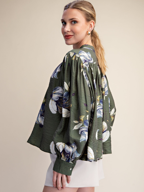 A woman wearing a dark olive green blouse with a blue, white, and yellow floral print, paired with white shorts, photographed against a solid beige studio background.