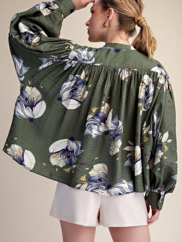 A woman wearing a sage green long-sleeve blouse with a large white and blue floral print, paired with white shorts, shown from a back-three-quarter view against a solid beige background.