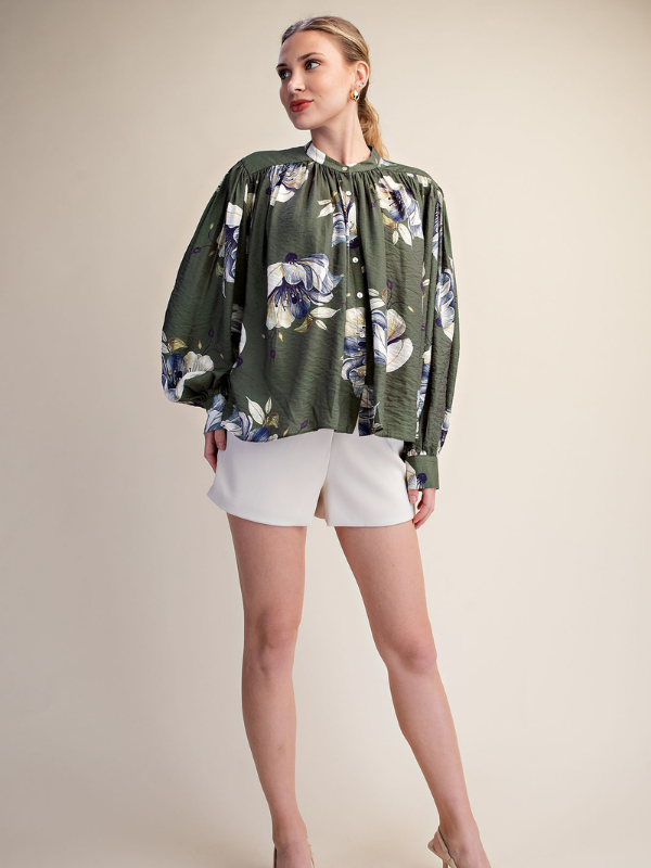 A woman modeling a green floral long-sleeve blouse with white shorts against a plain beige studio background.