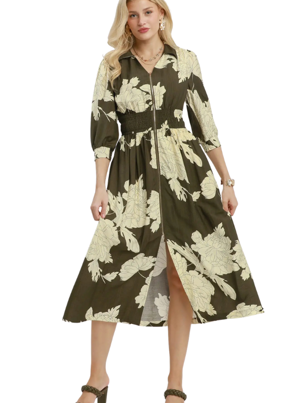 A woman modeling a mid-length olive green shirt dress with a large cream floral print, featuring a collar, three-quarter sleeves, a tie waist, and a front slit, set against a plain white background.