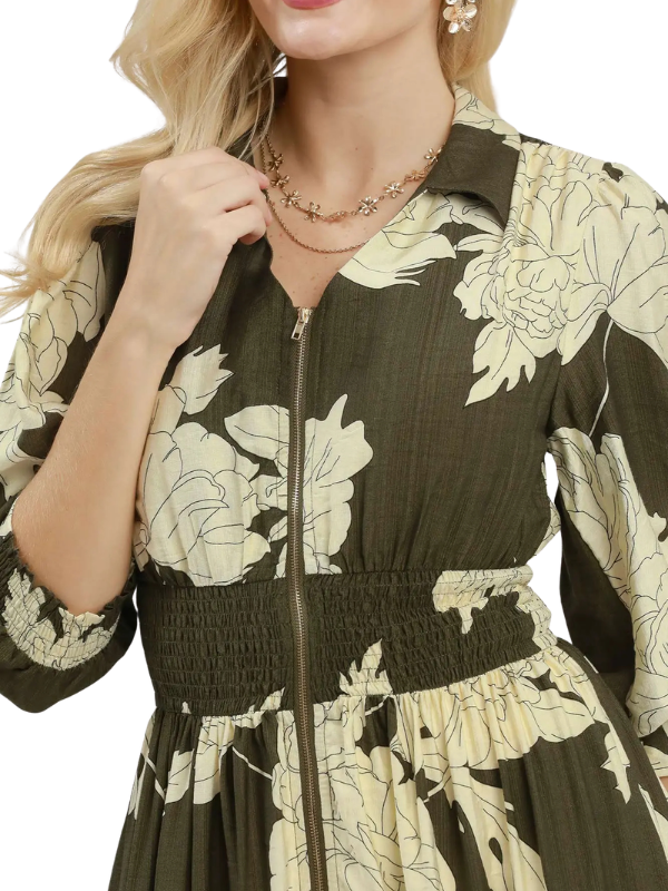 A close-up of a woman wearing a dark olive green and cream floral print dress with a smocked waist and a front zipper.