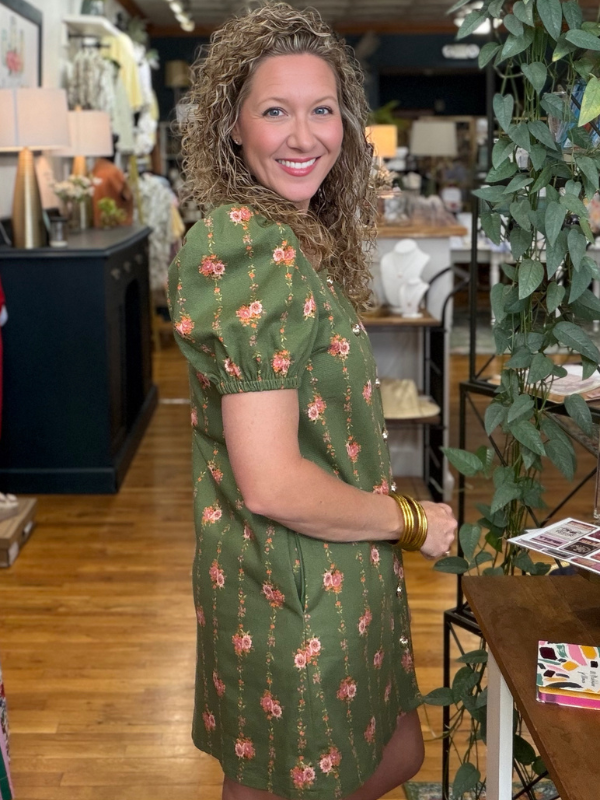 Woman in a green floral dress standing in a store with plants and furniture.