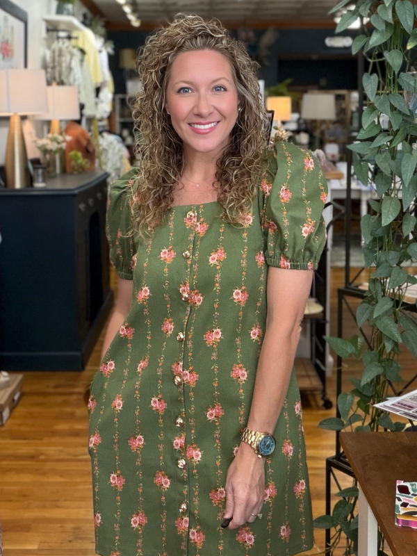 Woman in a green floral dress standing in a store with plants and furniture.