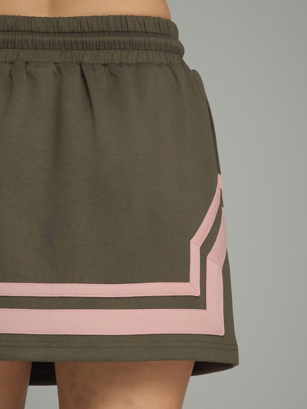 Close-up of a dark olive green athletic skirt with a pink geometric stripe detail along the side, worn by a model against a solid gray background.