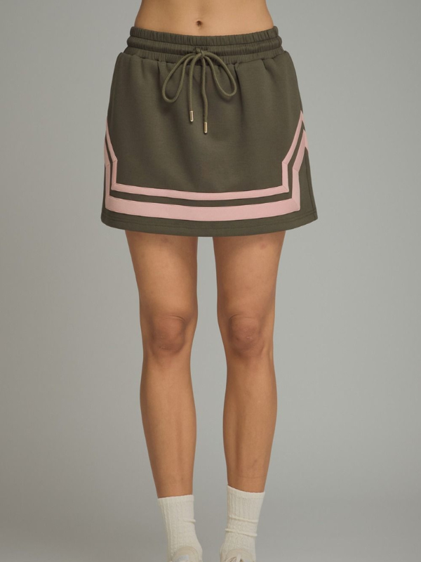 An olive green athletic mini skirt with pink geometric stripe accents and a matching drawstring waist, shown on a model against a neutral gray background.