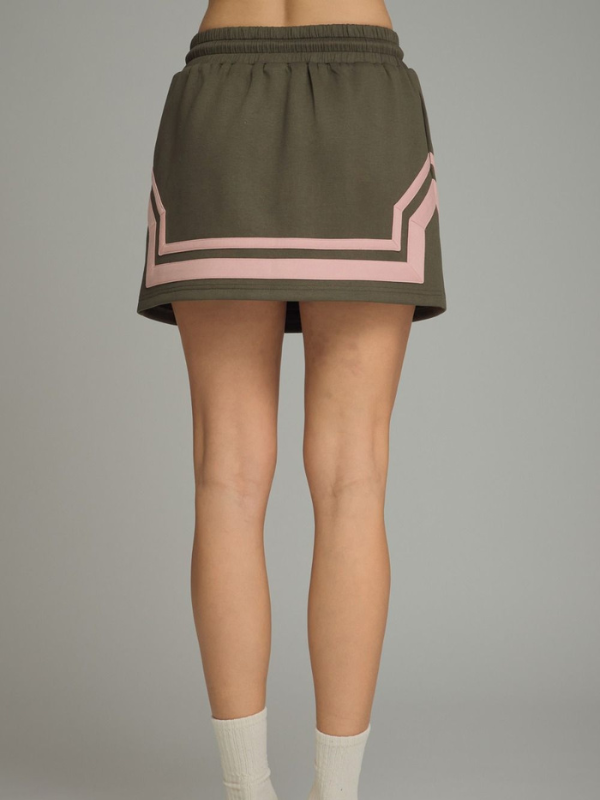 An olive green athletic mini skirt with a pink geometric stripe pattern near the hem, shown on a model from the waist down against a solid gray background.