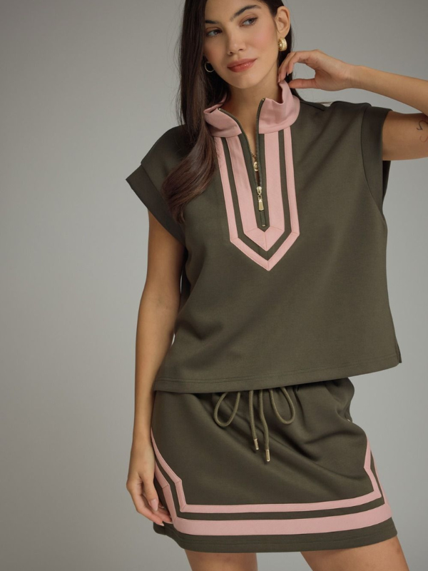 A woman modeling a matching olive green athletic set consisting of a short-sleeve crop top with a pink and dark green chevron zipper detail and a matching drawstring mini skirt with pink stripe accents, set against a solid gray studio background.