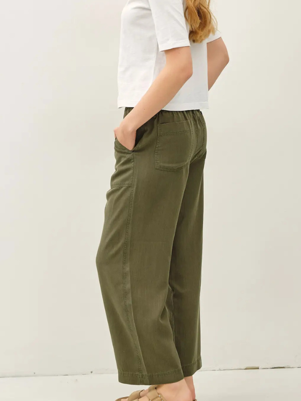 A person shown from the side wearing olive green wide-leg cropped pants with a white short-sleeve top and tan sandals, standing against a plain off-white wall.