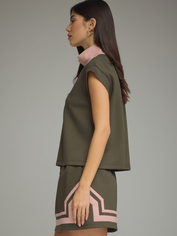 A woman shown in side profile wearing a sleeveless olive green top and matching skirt with light pink geometric trim, against a plain gray studio background.