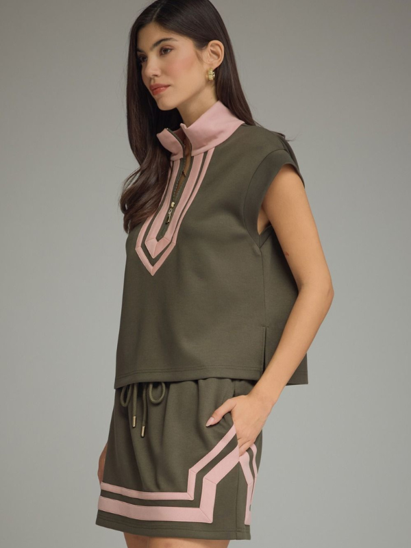A woman modeling a sleeveless olive green top and matching mini skirt set with light pink geometric trim details, photographed against a solid gray studio background.