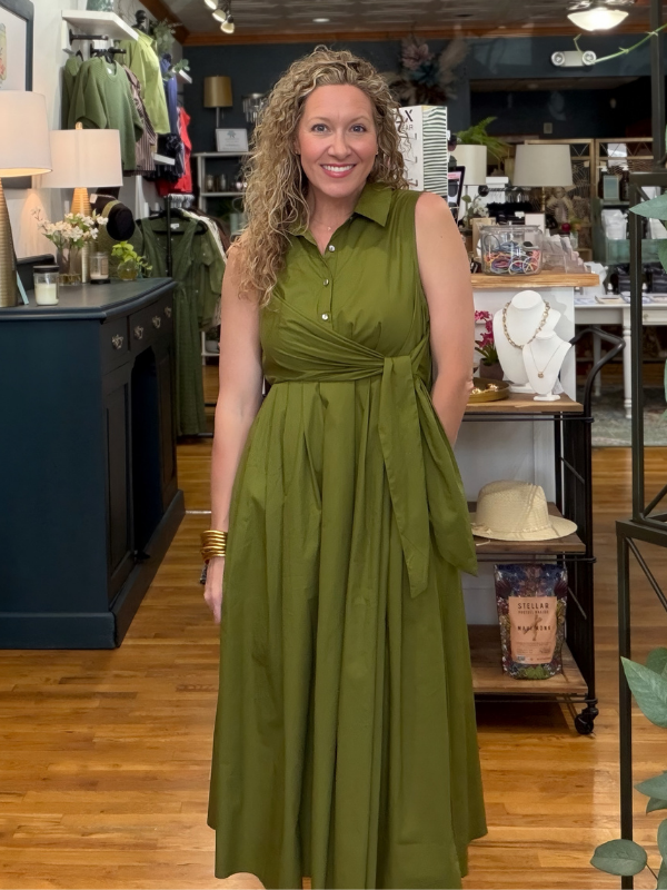 A woman with curly blonde hair stands in a boutique wearing a sleeveless olive green midi dress with a collared neckline, buttoned front, and a self-tie waist sash. The background shows a retail store interior with clothing racks, lamps, and wooden floors.