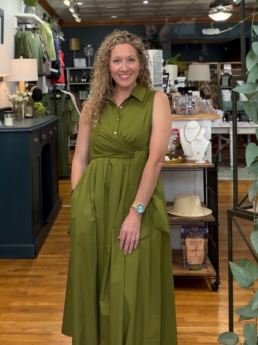 A woman with curly blonde hair wearing a sleeveless olive green midi dress with a button-down collar and gathered waist, standing in a retail boutique.