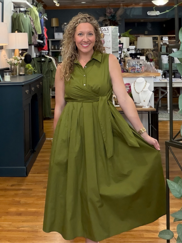 A woman modeling an olive green sleeveless midi dress with a collared neckline, buttoned front, and tied waist sash, standing inside a retail boutique.