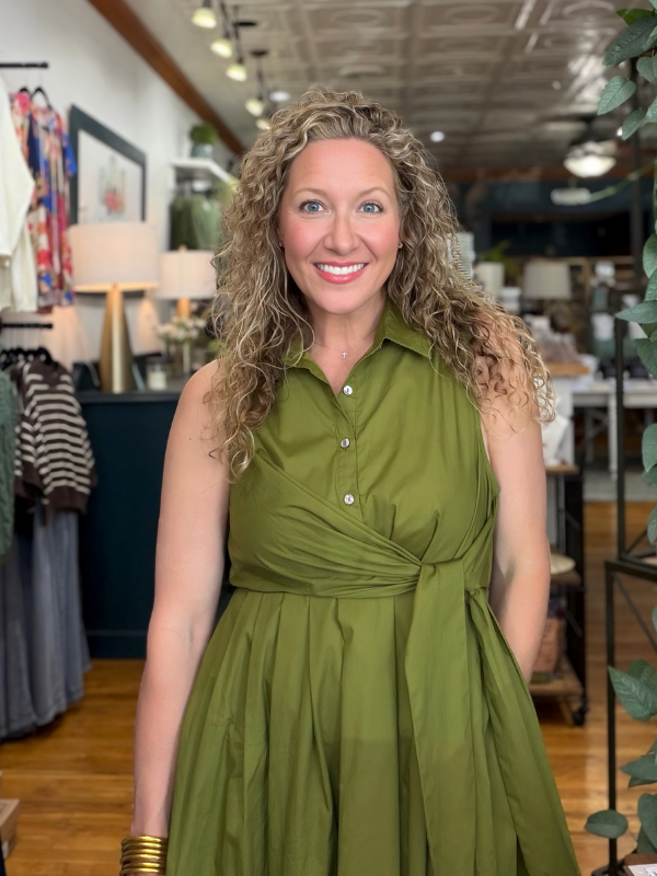 A smiling woman with curly blonde hair wearing an olive green sleeveless button-up dress with a twisted waist detail, standing inside a retail boutique.