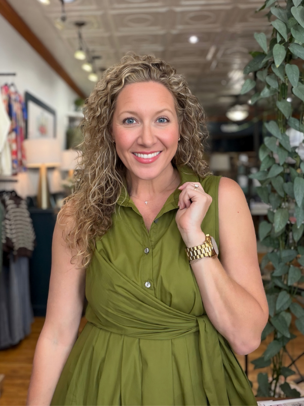 A smiling woman with curly blonde hair wearing a sleeveless olive green wrap-style dress, standing inside a boutique with store fixtures and plants in the background.