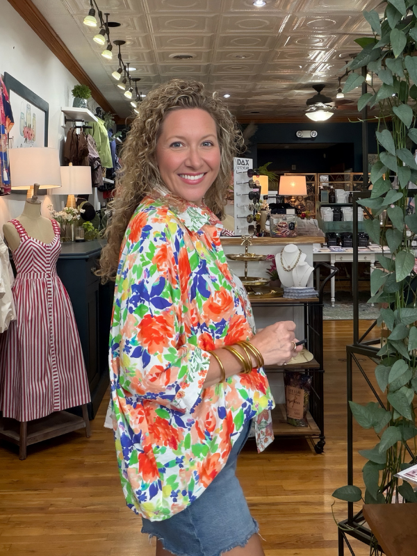 A smiling woman with curly hair stands in a boutique wearing a colorful floral long-sleeve blouse with blue denim shorts. The blouse features a vibrant pattern of orange, red, green, and blue flowers on a white background.