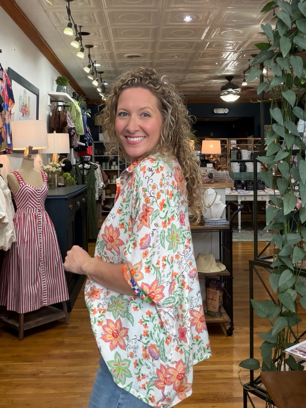 A woman with curly hair smiling and looking back over her shoulder inside a clothing boutique. She is wearing a loose white button-up shirt with a colorful floral print in orange, pink, and green, paired with blue jeans.