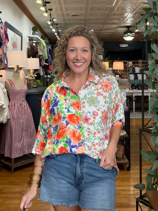 A smiling woman with curly blonde hair stands inside a boutique wearing a white short-sleeve button-up shirt with a vibrant orange, red, and blue floral print, paired with blue denim shorts.