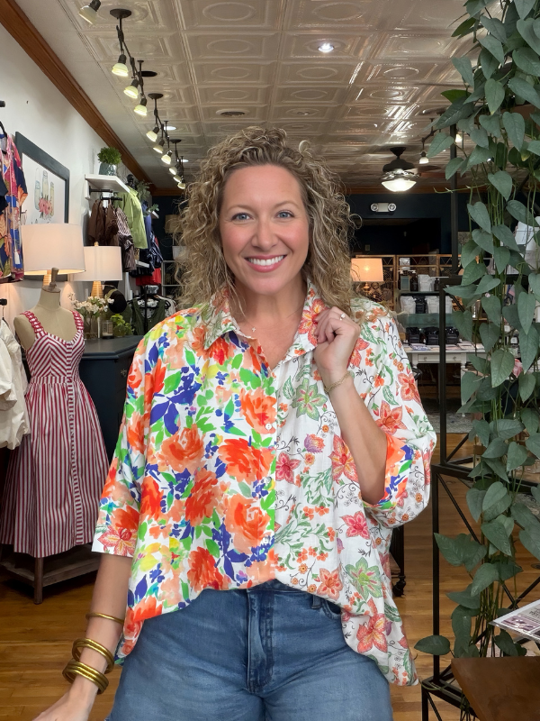 A smiling woman with curly blonde hair stands inside a clothing boutique wearing a white button-up shirt with a vibrant, multicolor floral print in orange, green, blue, and red, paired with blue jeans.