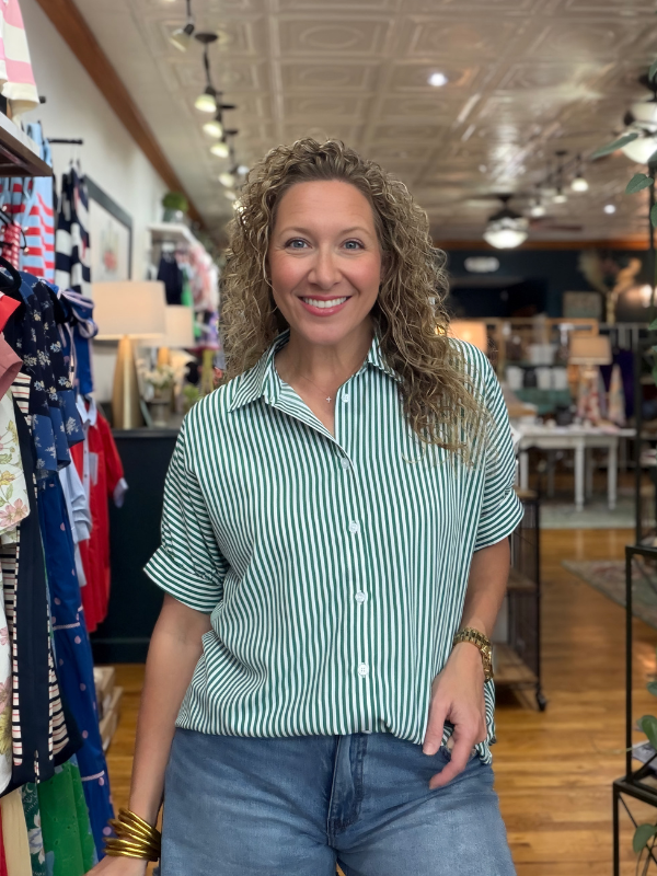 Woman in a striped shirt standing in a store with clothing racks and products in the background.