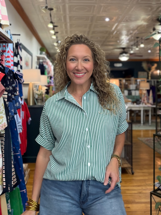 Woman in a striped shirt standing in a store with clothing racks and products in the background.