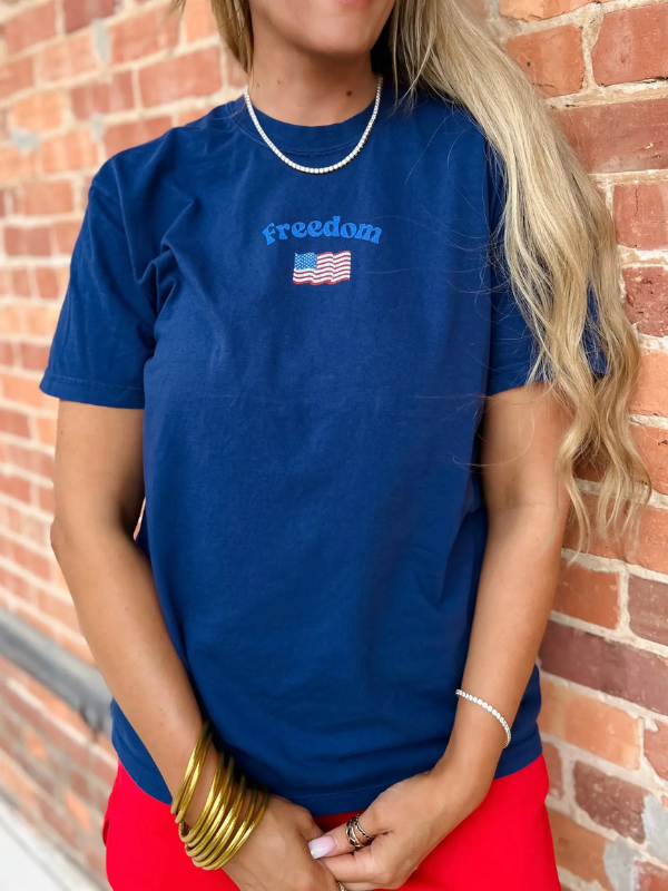 Person wearing a blue t-shirt with 'freedom' and an American flag design, standing against a brick wall.