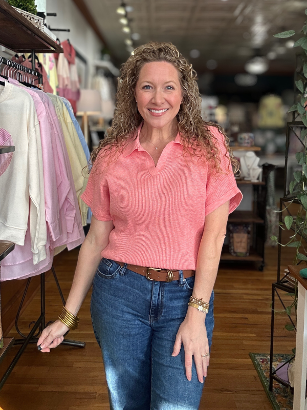 Woman in a pink shirt and blue jeans standing in a store with clothing racks in the background.