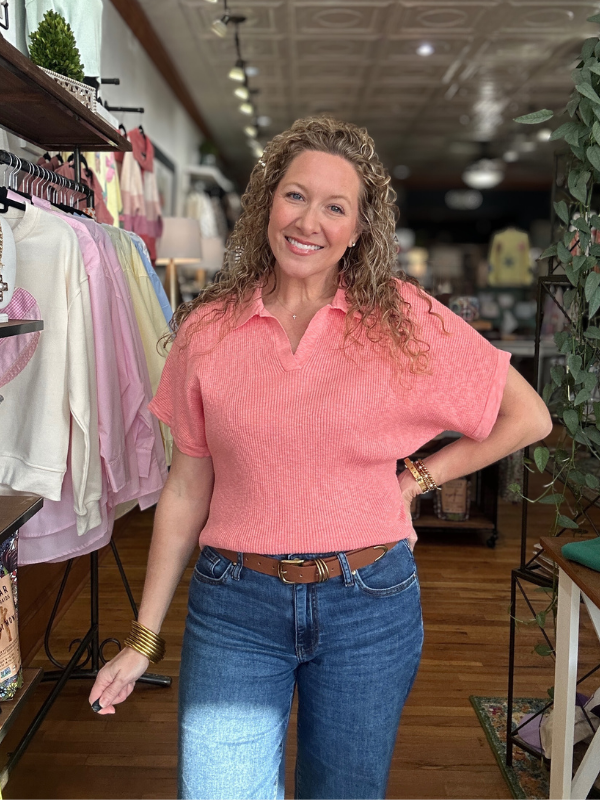 Woman in a pink shirt and blue jeans standing in a store with clothing racks in the background.