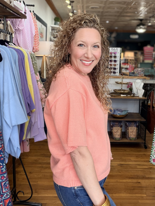 Woman in a pink sweater standing in a store with clothing racks and products in the background.