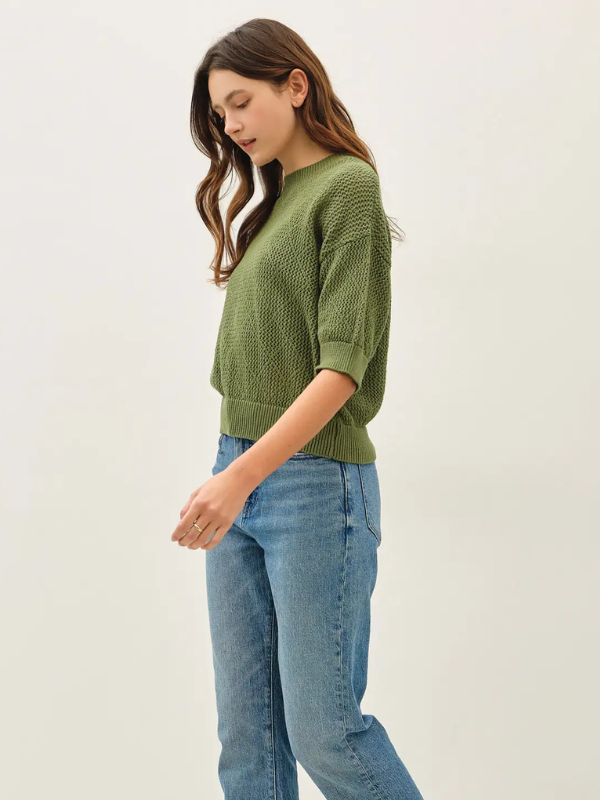 A woman wearing a textured olive green knit sweater and light blue jeans, standing against a plain off-white studio background.