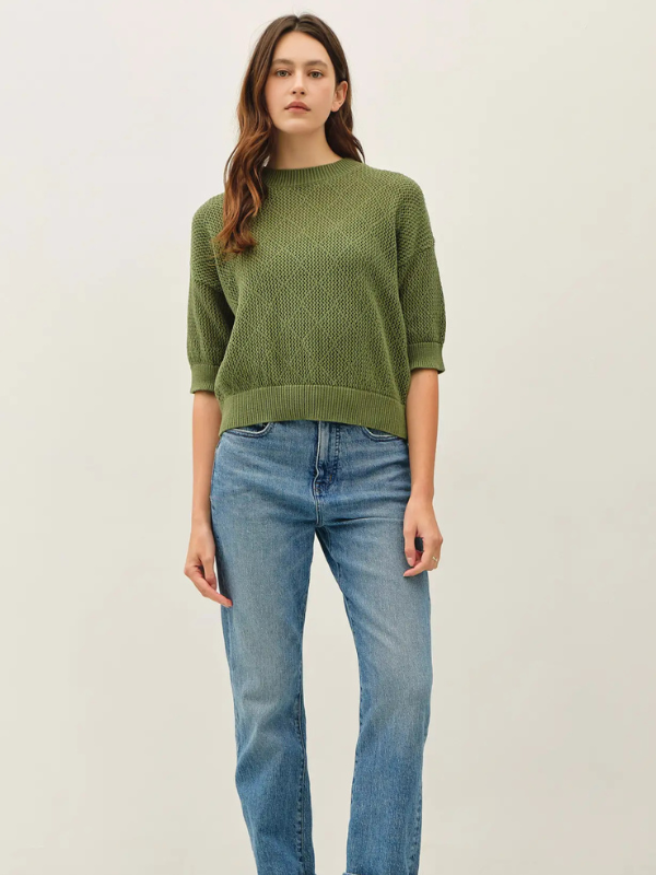 A woman modeling a textured olive green knit sweater with three-quarter sleeves, paired with light-wash blue jeans, against a plain off-white studio background.