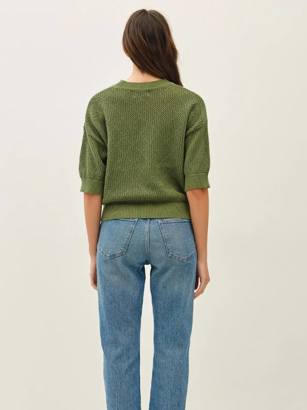 A woman modeling a textured olive green knit sweater with three-quarter sleeves, paired with light-wash blue jeans, against a plain off-white studio background.